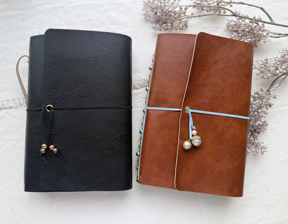 Black and brown leather refillable journals with beaded closures, next to dried flowers
