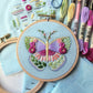 Finished butterfly sampler embroidery in purple, pink and green on light blue fabric, with the stitch guide and floss