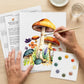 Hands painting the mushroom watercolor outline with the kit's brush and paint card