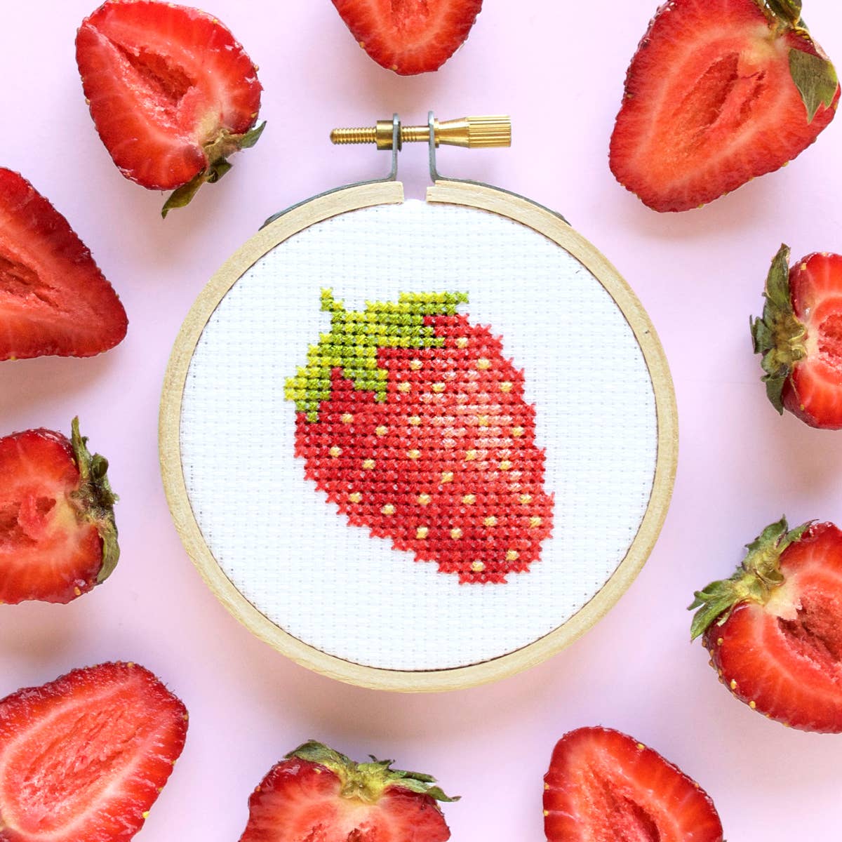 Strawberry cross stitch hoop surrounded by sliced strawberries on pink