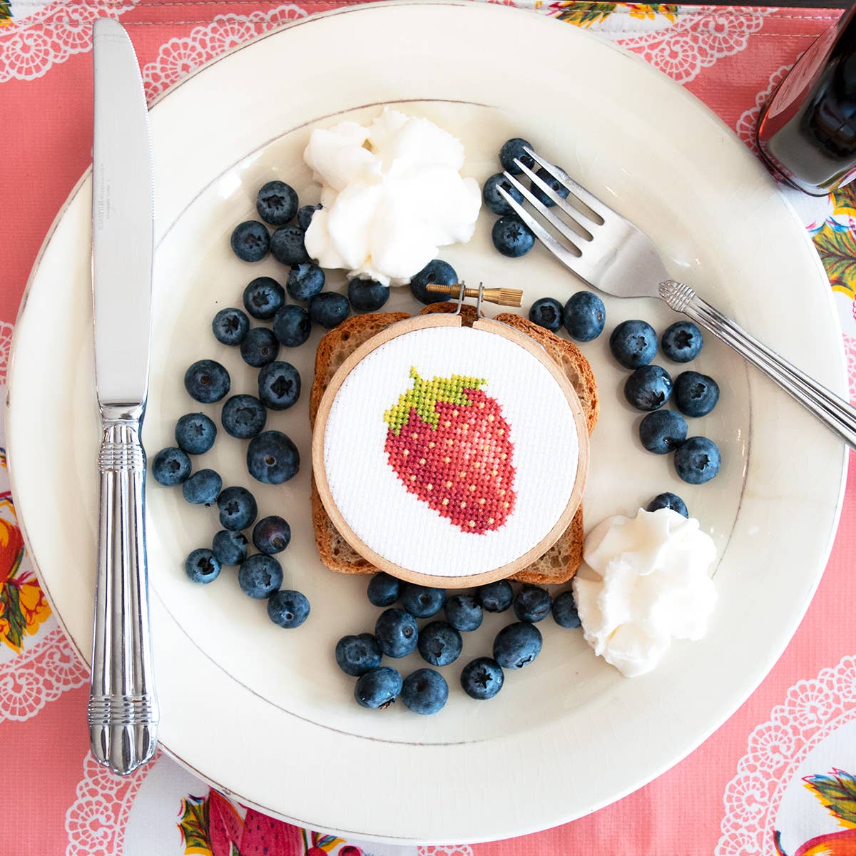 Strawberry cross stitch hoop on toast with blueberries and whipped cream on a white plate