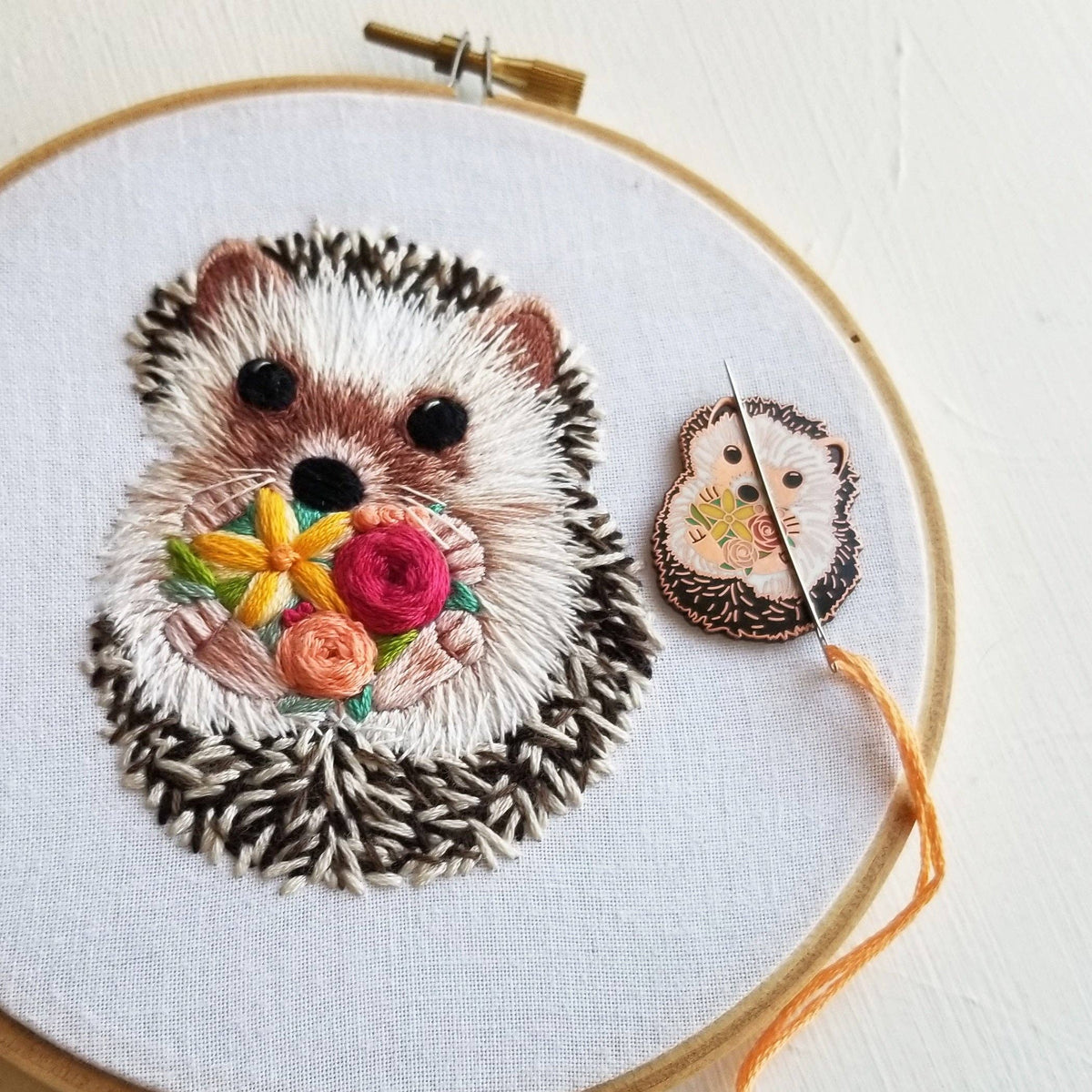 Embroidered hedgehog hoop beside the needle minder holding a needle and orange floss