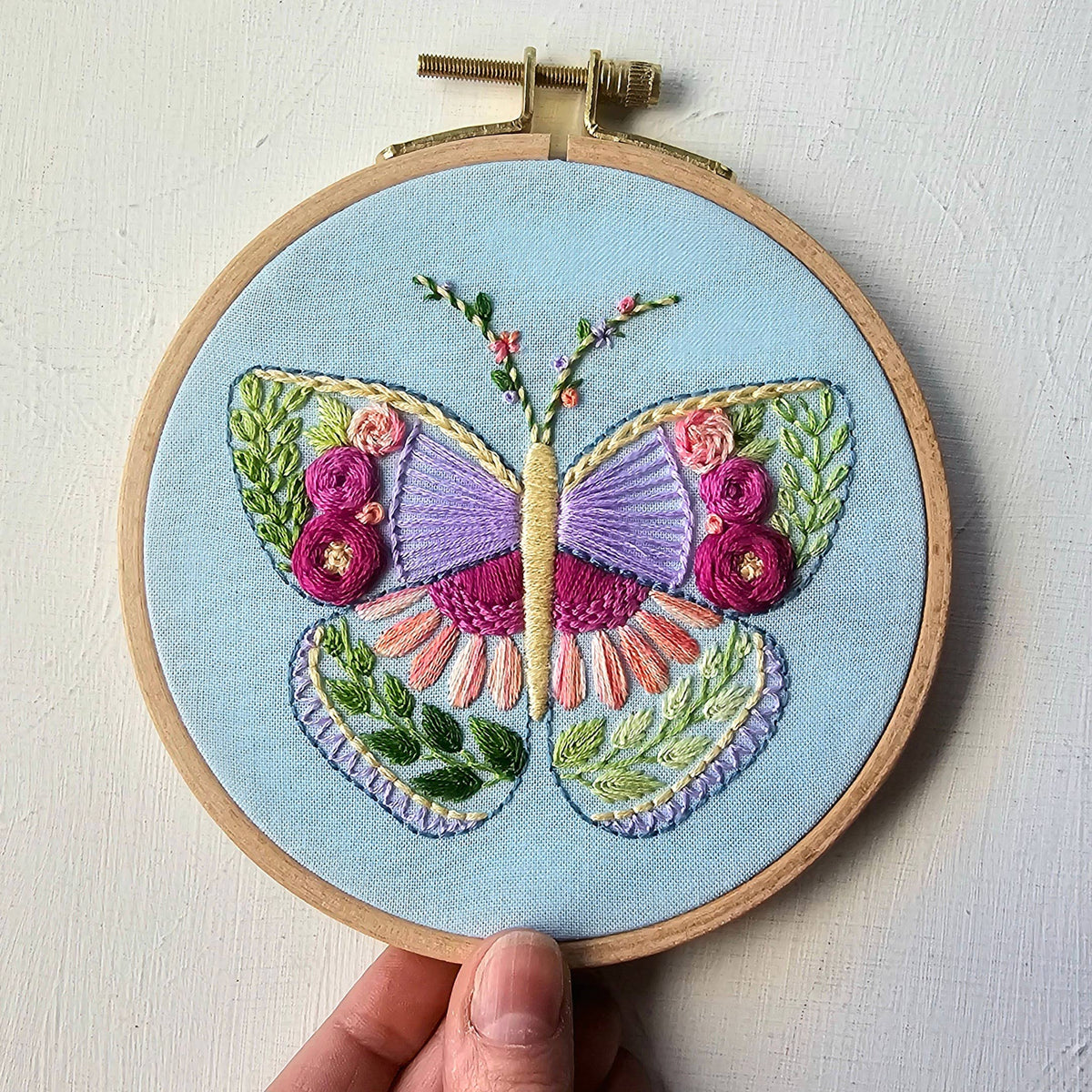 Hand holding the finished butterfly sampler in a wooden hoop