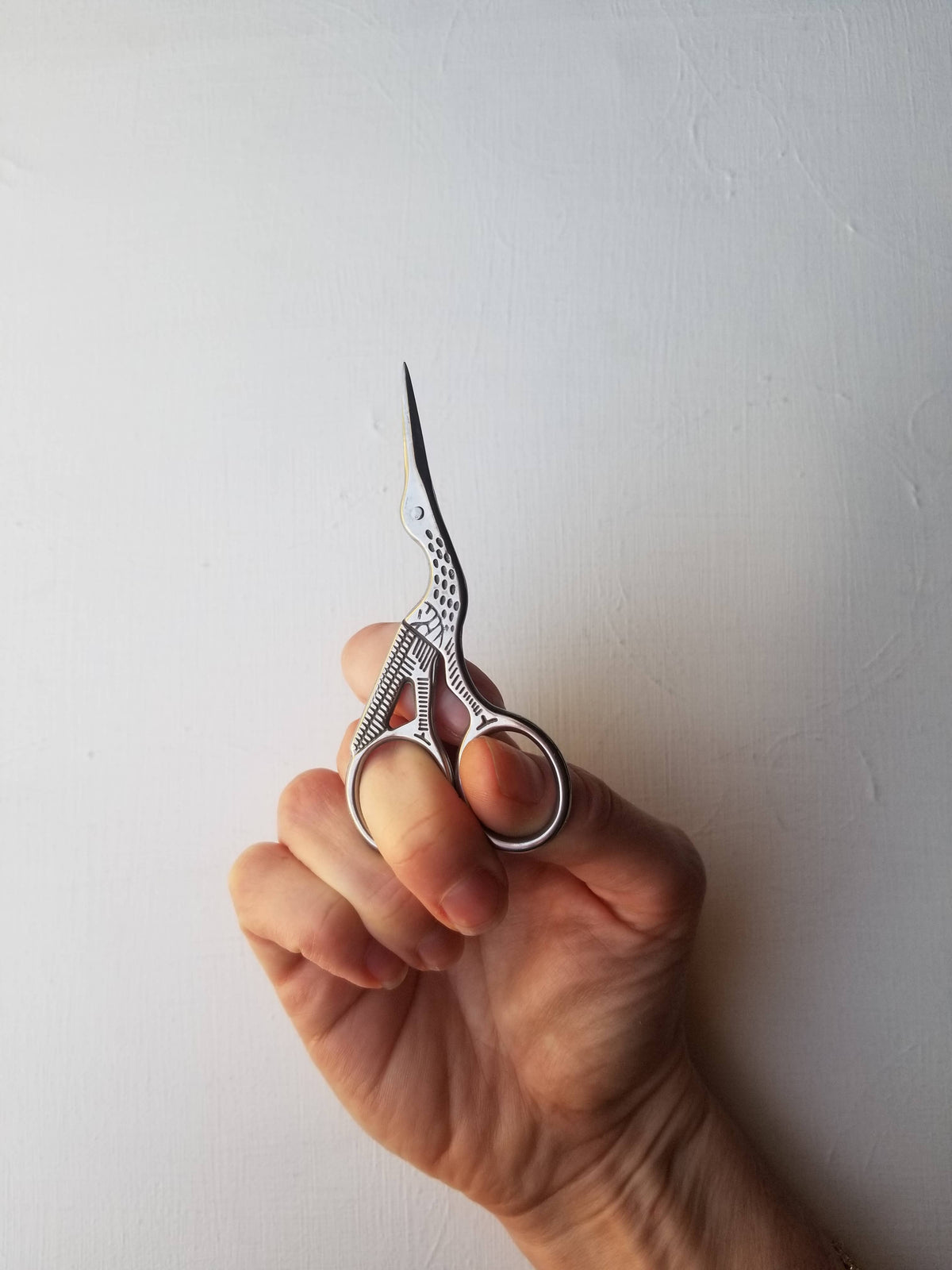 Hand holding the stork embroidery scissors upright against a white wall