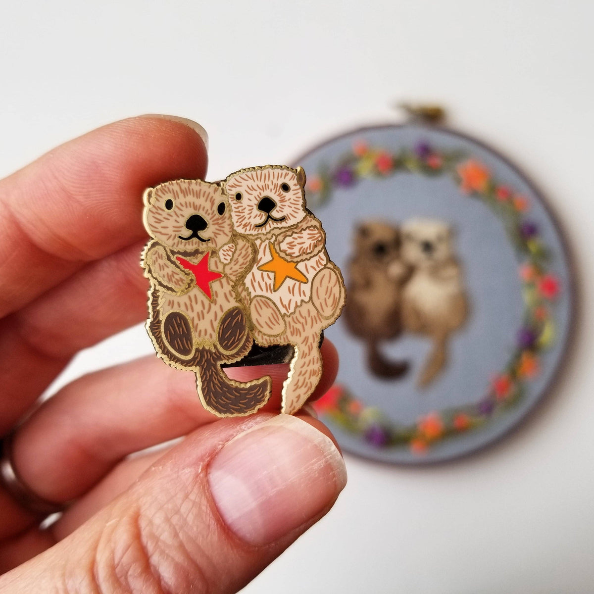 Hand holding a sea otter couple enamel needle minder in front of a matching embroidery hoop