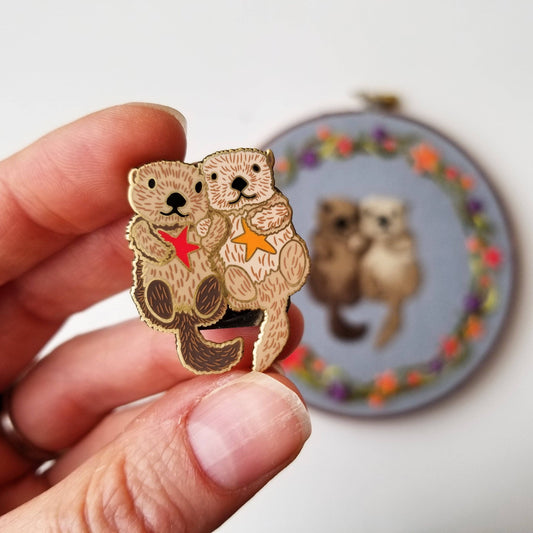Hand holding a sea otter couple enamel needle minder in front of a matching embroidery hoop