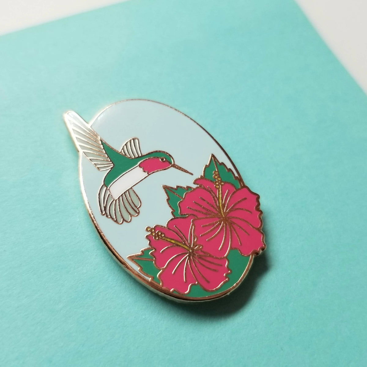 Oval enamel needle minder with a hummingbird above pink hibiscus flowers