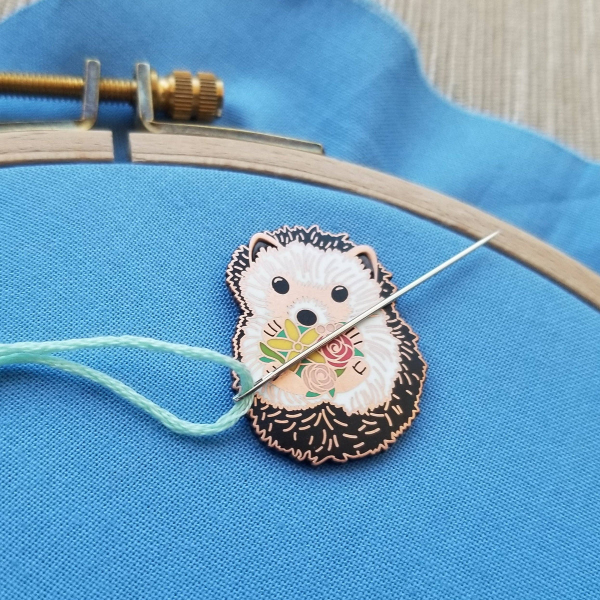 Hedgehog needle minder holding a threaded needle on blue fabric in an embroidery hoop