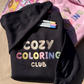Black and pink Cozy Coloring Club crewnecks with pastel embroidered lettering, markers and a coloring book