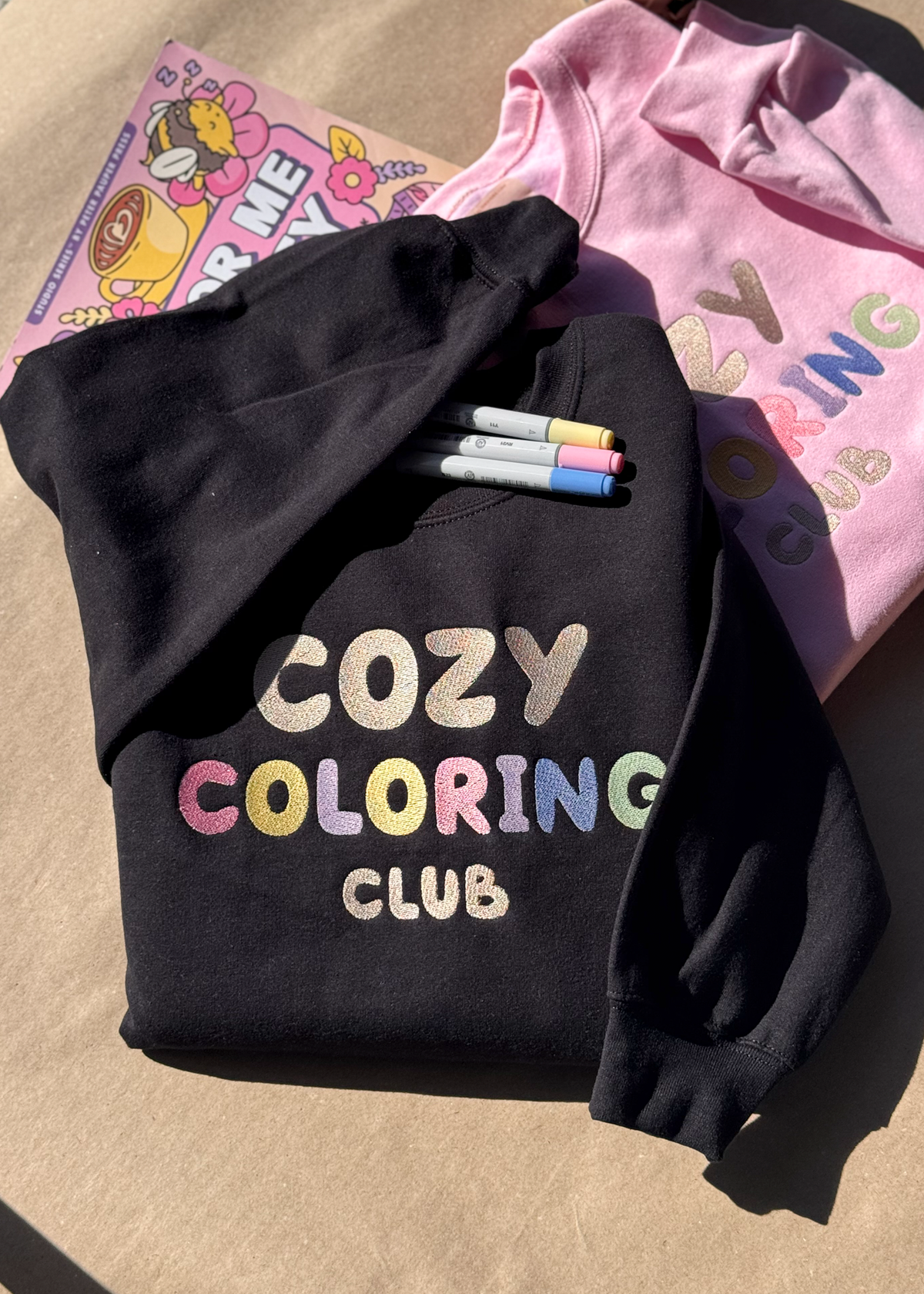 Black and pink Cozy Coloring Club crewnecks with pastel embroidered lettering, markers and a coloring book
