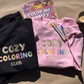 Black and pink Cozy Coloring Club crewnecks laid flat with markers, a coffee cup and a coloring book