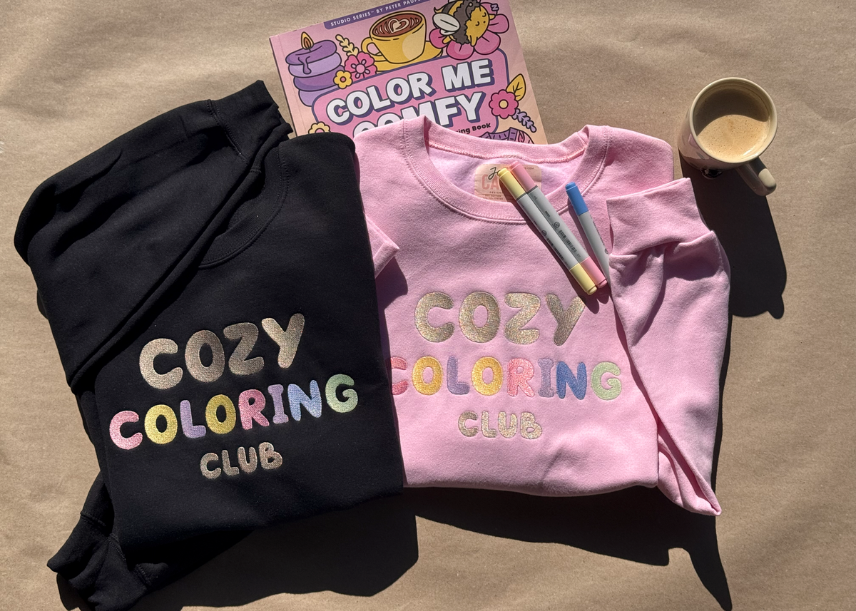 Black and pink Cozy Coloring Club crewnecks laid flat with markers, a coffee cup and a coloring book