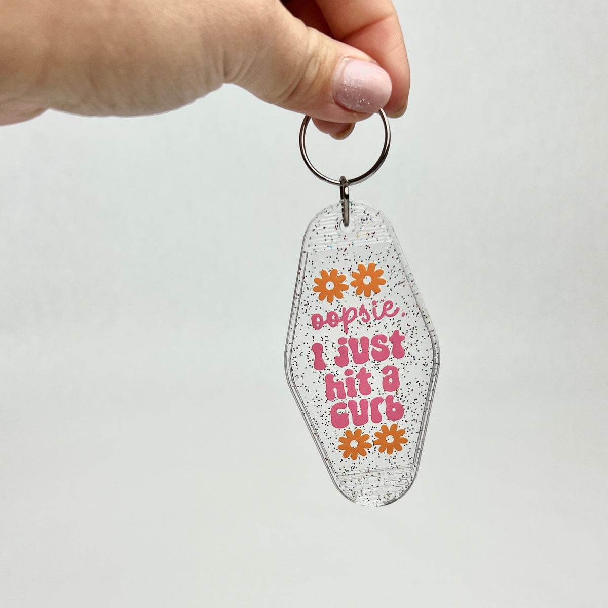 Hand holding a clear glitter motel-style keychain reading "oopsie, I just hit a curb"
