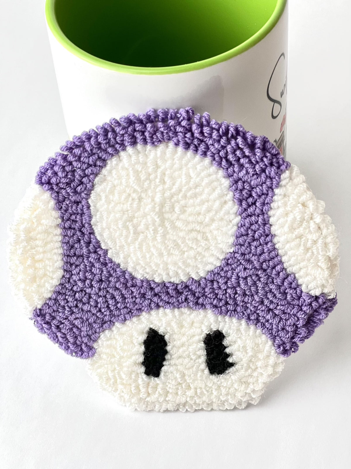 Purple and white punch needle mushroom coaster leaning against a mug