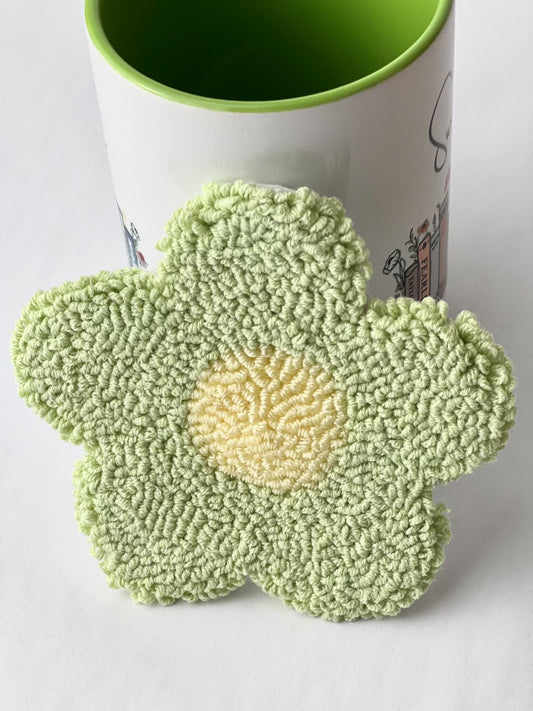 Green punch needle daisy coaster with a pale yellow center, leaning against a mug