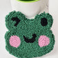 Green punch needle winking frog face coaster with pink cheeks, leaning against a mug