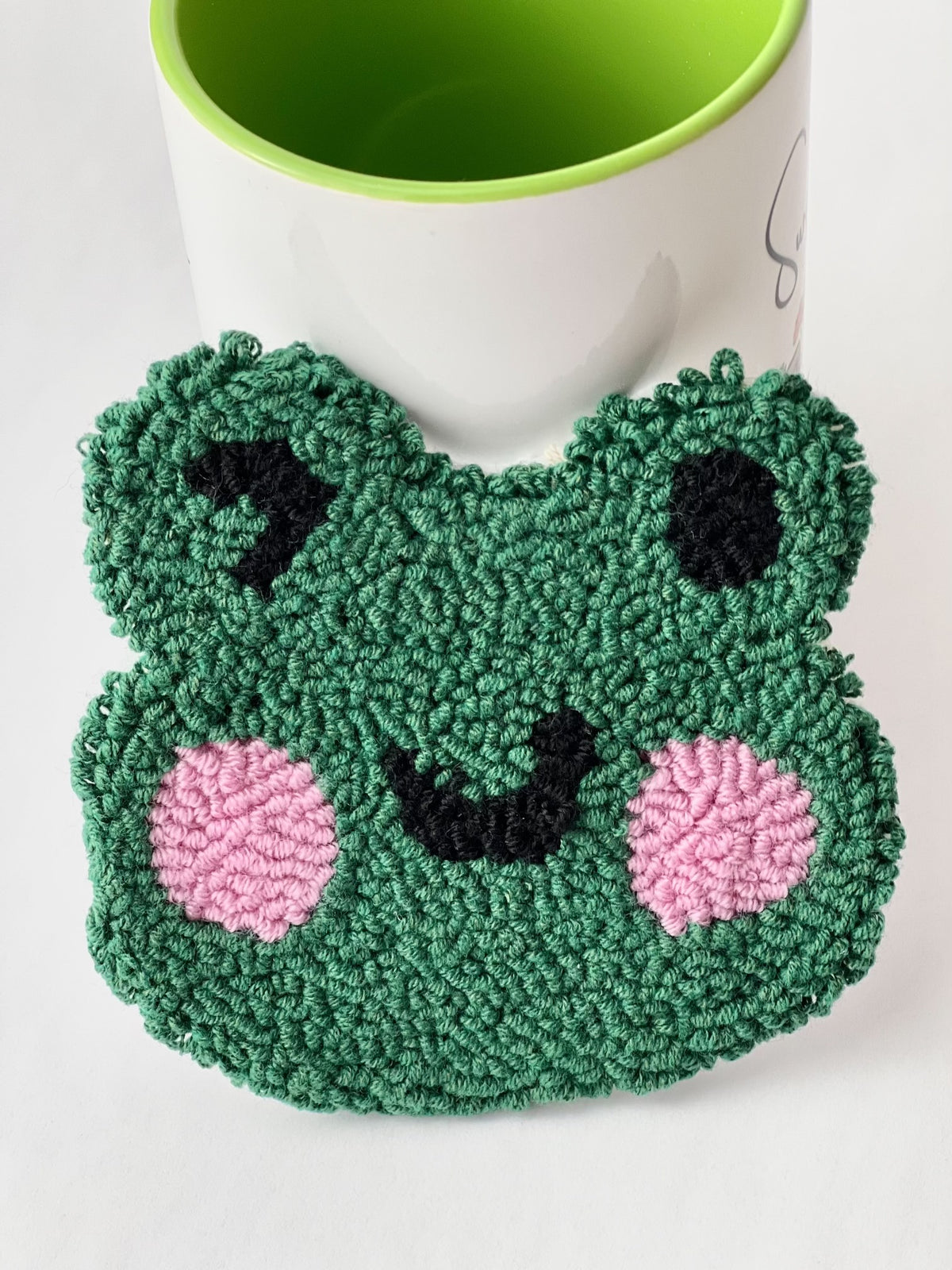 Green punch needle winking frog face coaster with pink cheeks, leaning against a mug