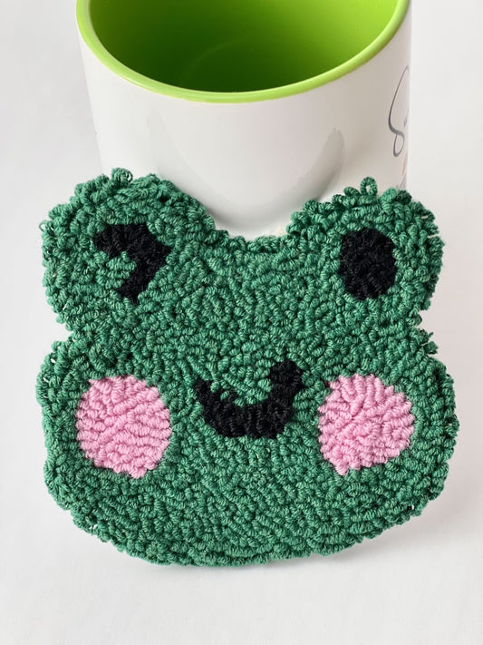 Green punch needle winking frog face coaster with pink cheeks, leaning against a mug