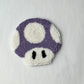 Purple and white punch needle mushroom coaster with black eyes on a white background