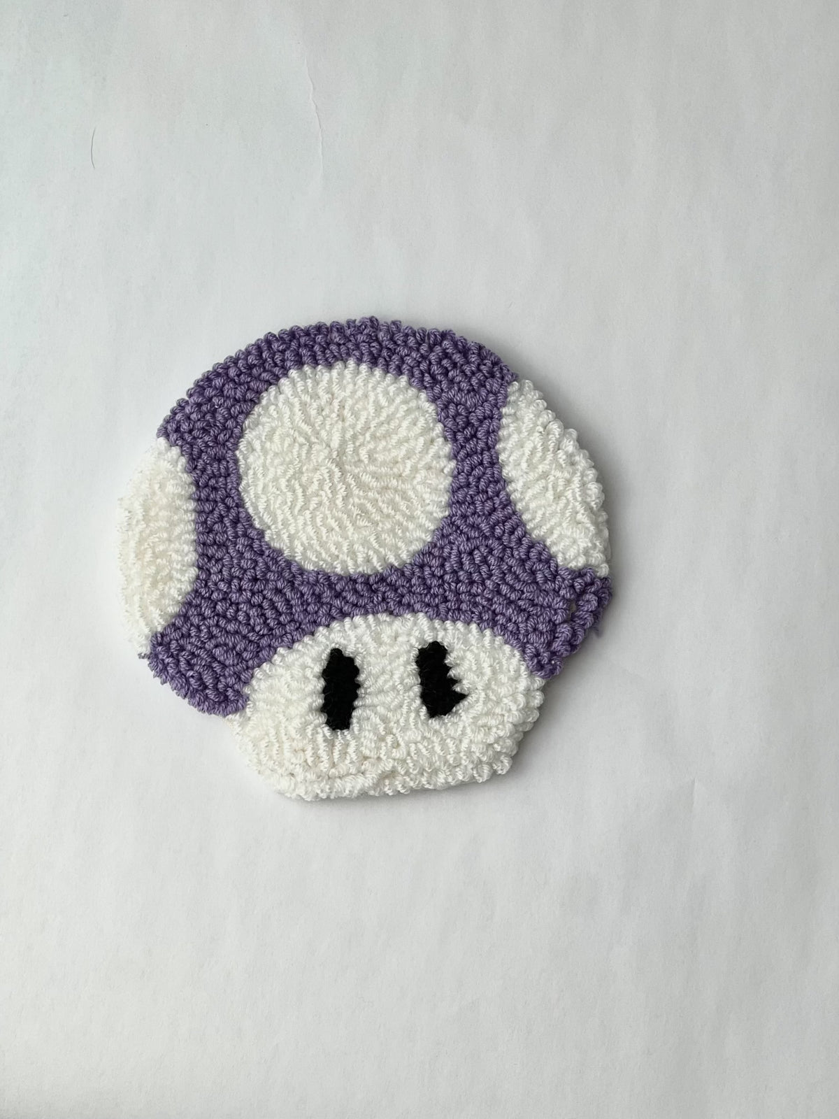 Purple and white punch needle mushroom coaster with black eyes on a white background