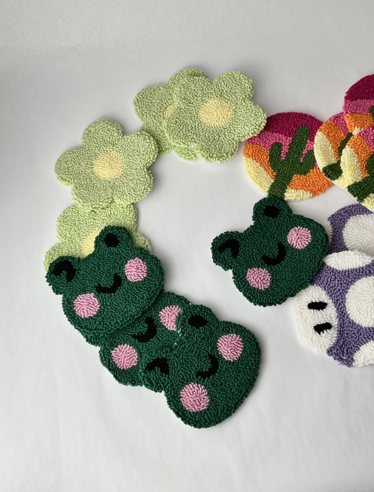 Stacks of punch needle coasters: winking frogs, green daisies, cactus sunsets and a purple mushroom