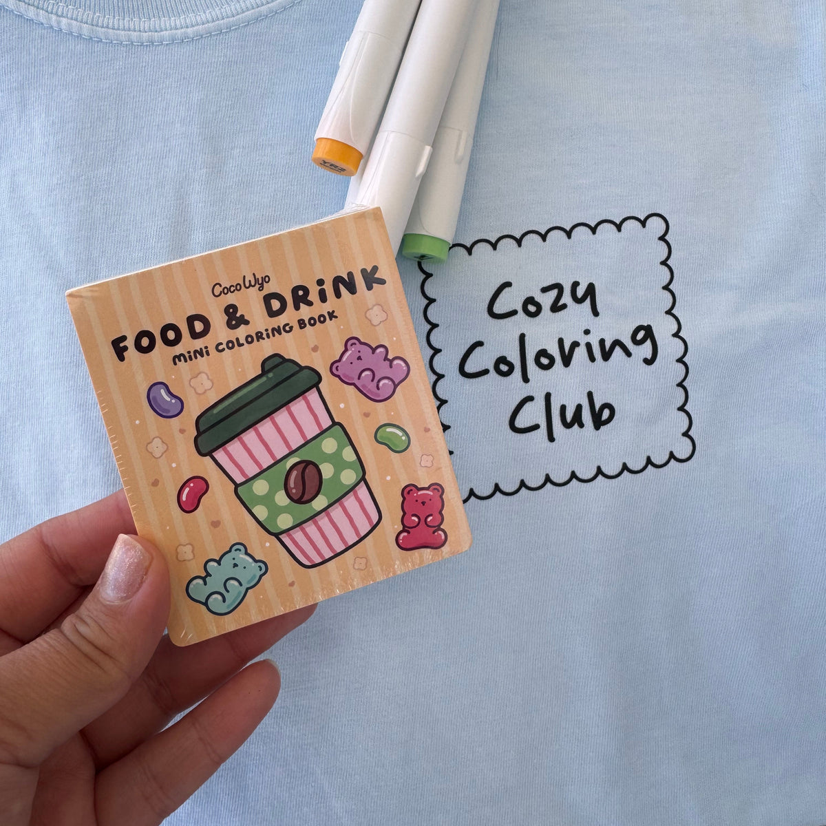 Hand holding the Food & Drink mini coloring book beside markers and a Cozy Coloring Club tee