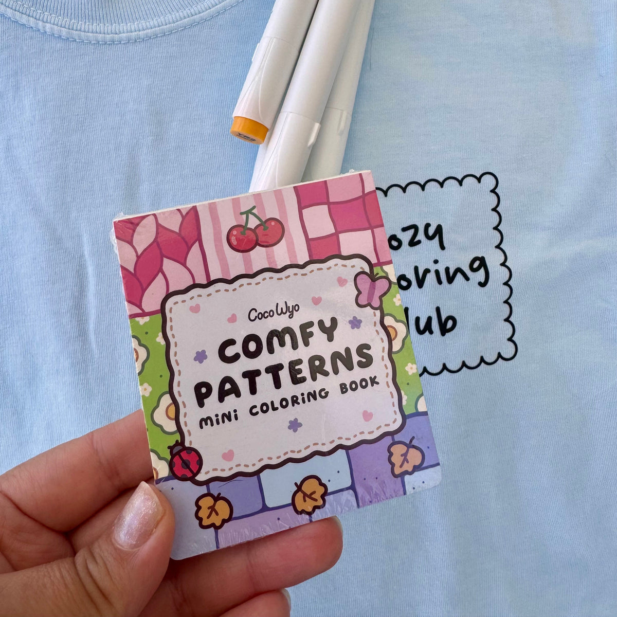 Hand holding the Comfy Patterns mini coloring book by Coco Wyo in front of markers and a blue tee