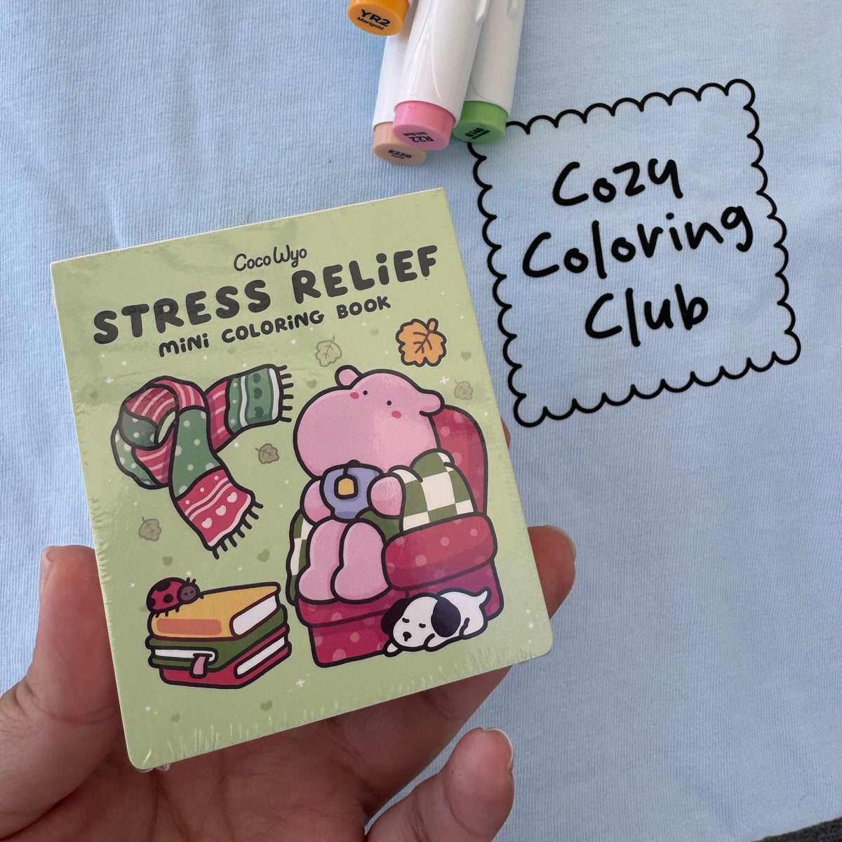 Hand holding the Stress Relief mini coloring book by Coco Wyo beside markers and a Cozy Coloring Club tee