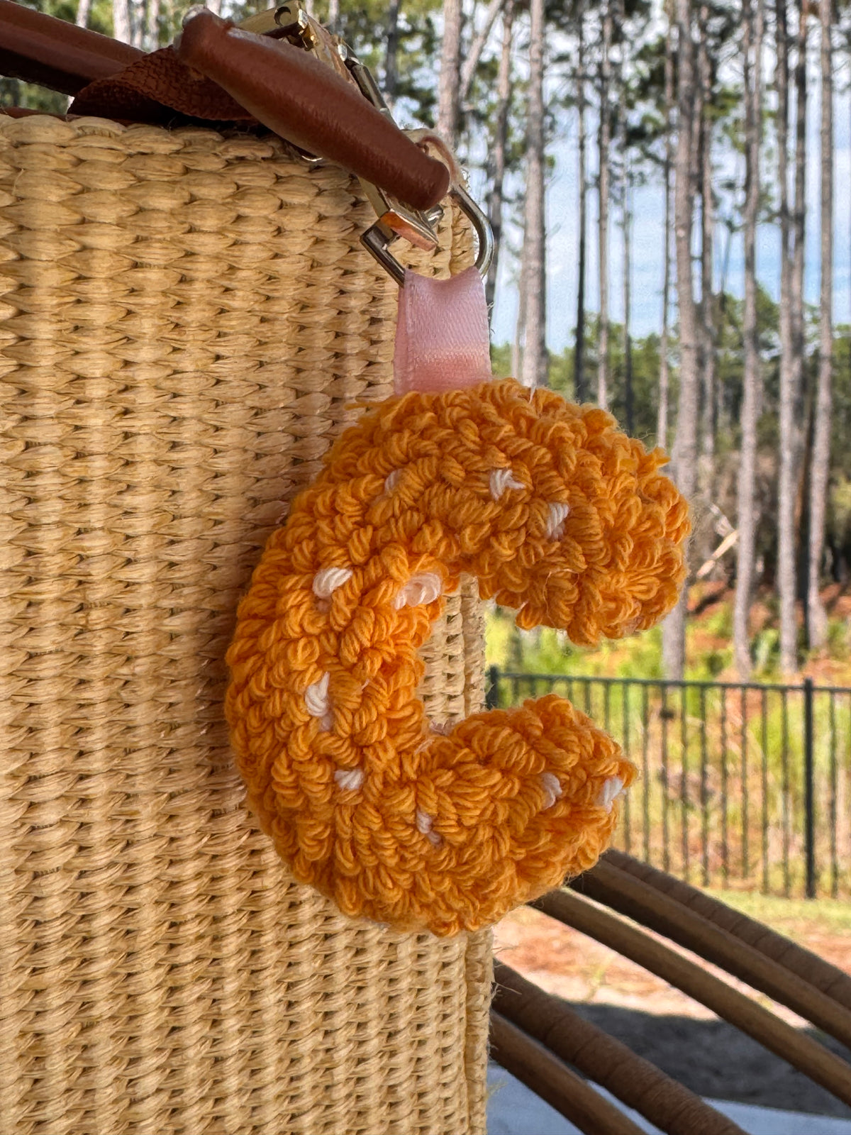 Orange punch needle letter C bag charm with white speckles, clipped to a woven straw bag
