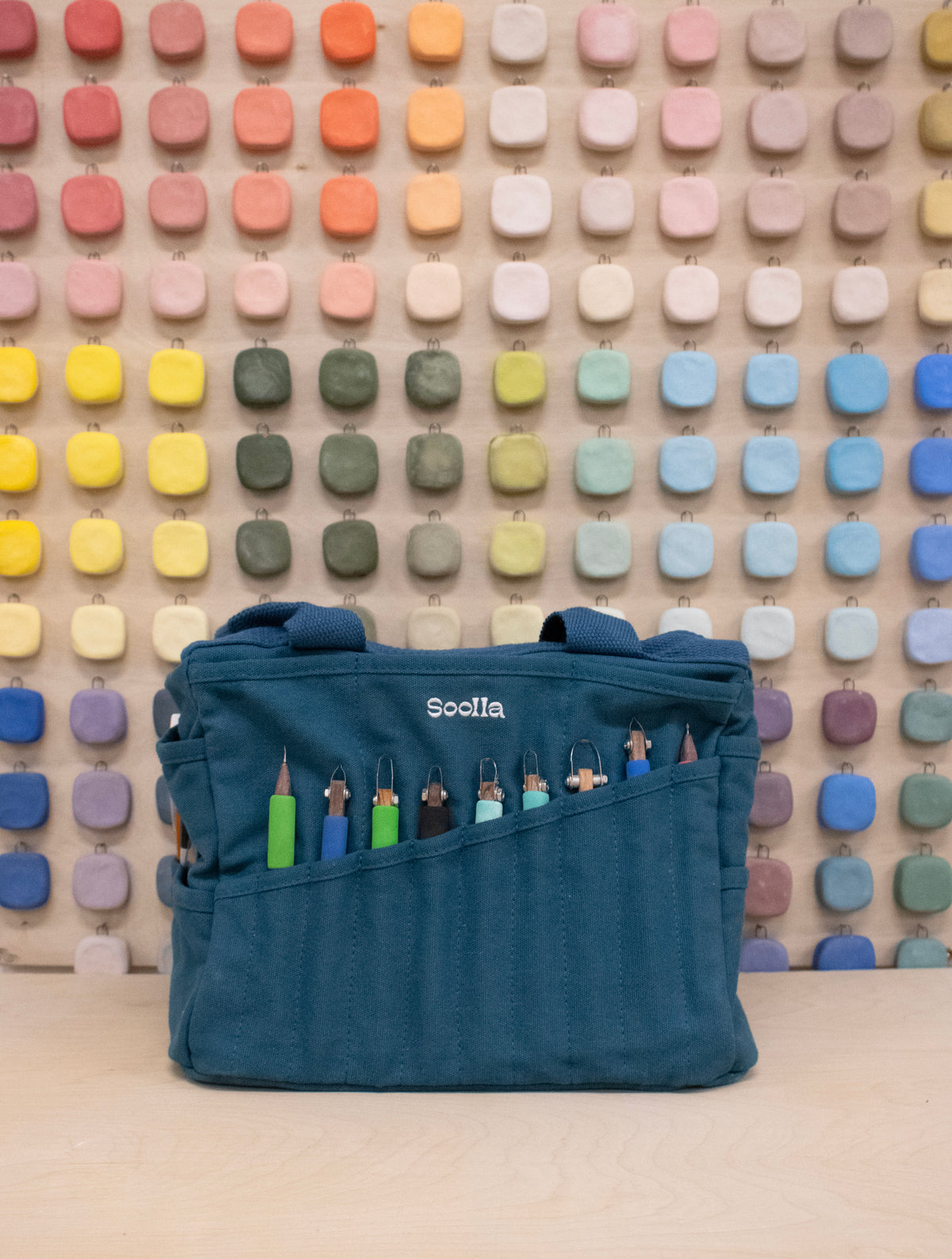 Craft bag with carving tools in the front pockets, in front of a wall of color swatches