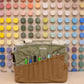 Sage and brown Soolla craft bag with carving tools in its pockets, in front of a wall of color swatches
