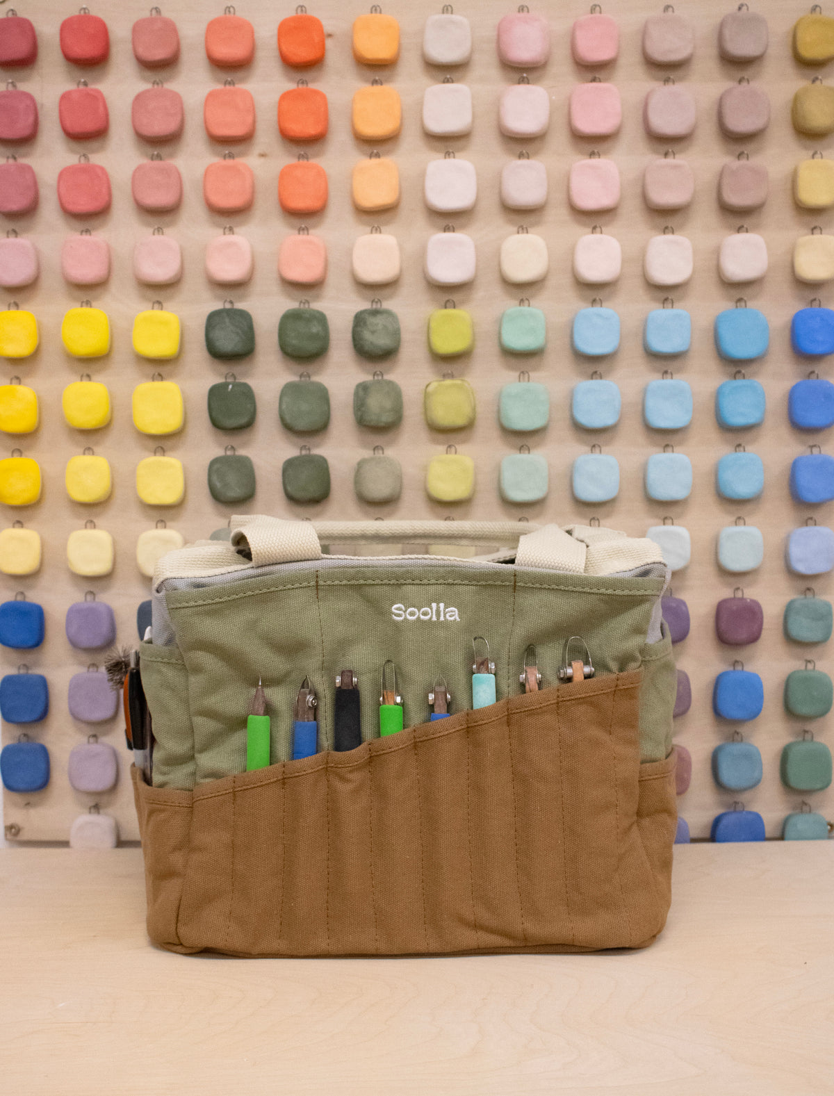Sage and brown Soolla craft bag with carving tools in its pockets, in front of a wall of color swatches