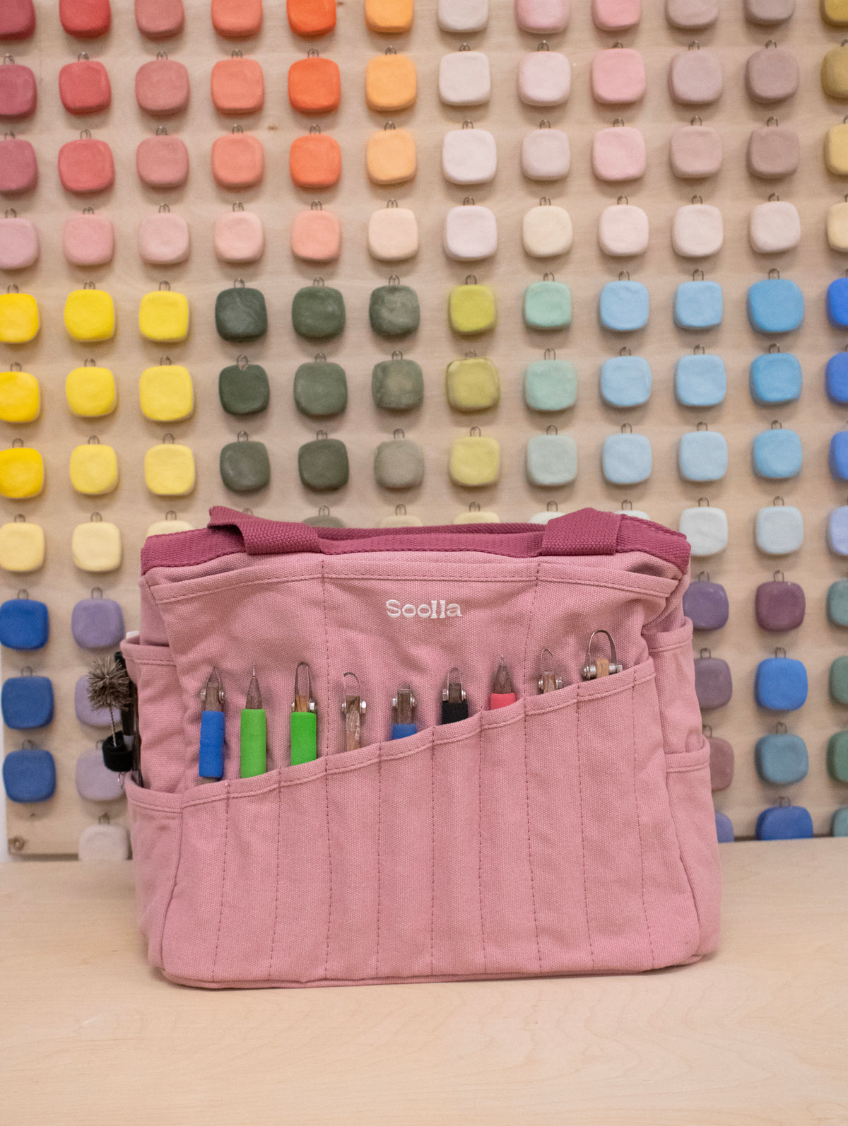 Dusty pink Soolla craft bag with carving tools in its pockets, in front of a wall of color swatches