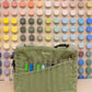 Green Soolla craft bag with carving tools in its pockets, in front of a wall of color swatches