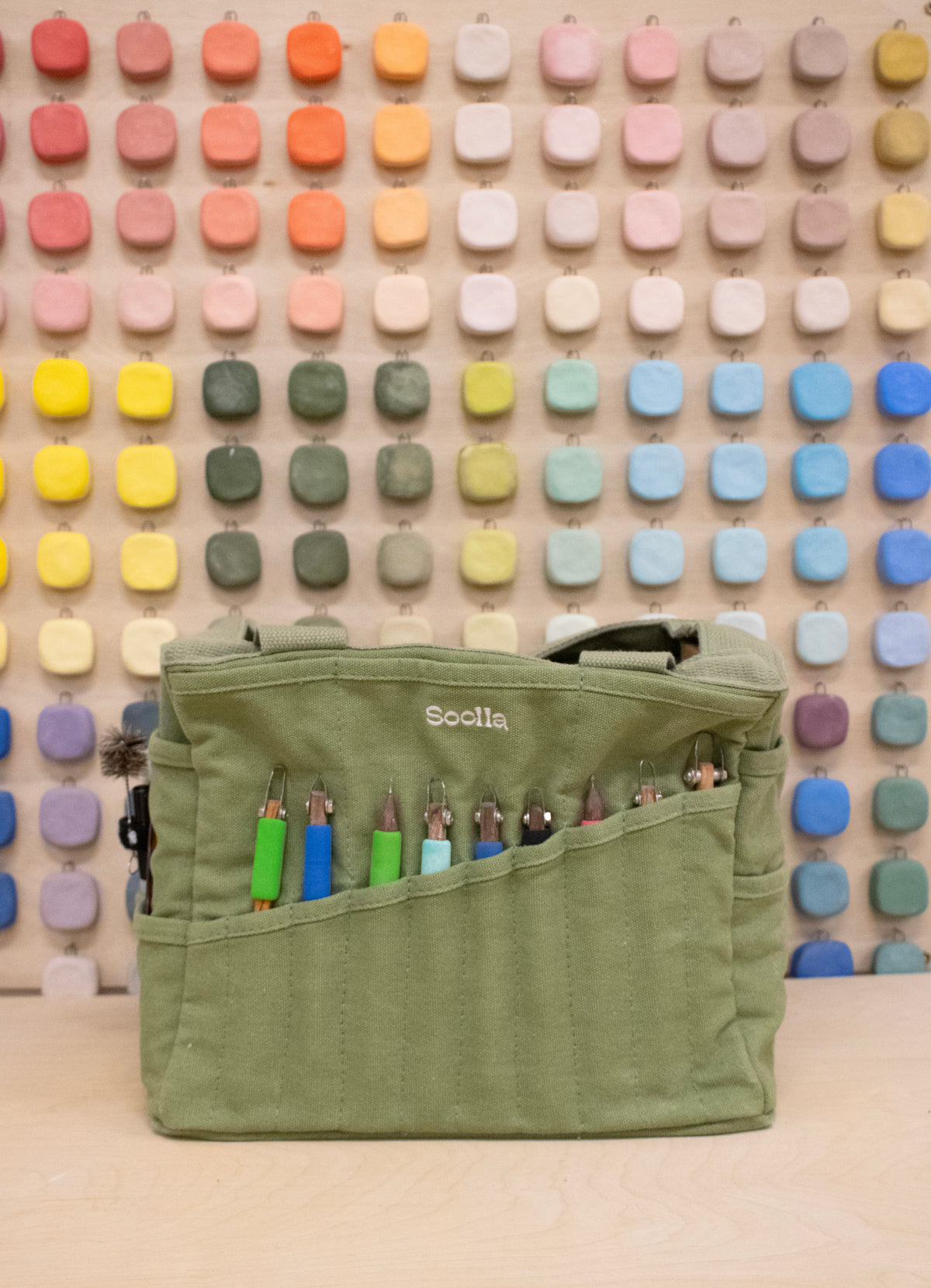 Green Soolla craft bag with carving tools in its pockets, in front of a wall of color swatches