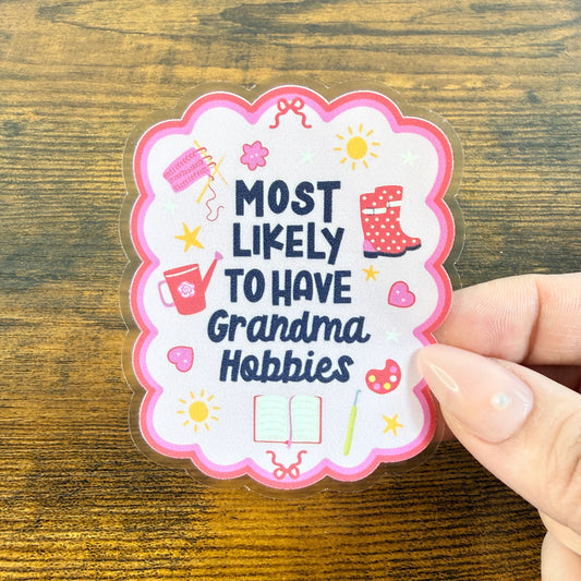 Hand holding a scalloped sticker reading "Most likely to have Grandma Hobbies" with knitting, rain boots and a watering can