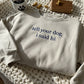 Sand crewneck embroidered "tell your dog i said hi" in navy, folded on a cream crochet blanket beside a paw-print mug and dog treats