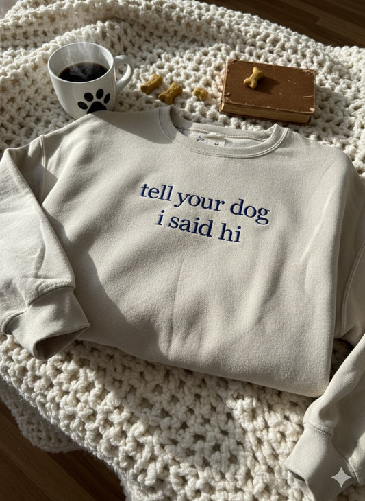 Sand crewneck embroidered "tell your dog i said hi" in navy, folded on a cream crochet blanket beside a paw-print mug and dog treats