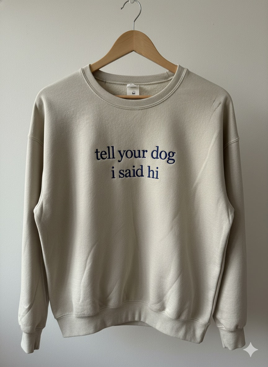 Sand "tell your dog i said hi" crewneck with navy embroidery on a wooden hanger