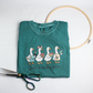 Folded teal Hobby Goose tee with four crafty geese and "just a crafty goose", beside scissors and an embroidery hoop