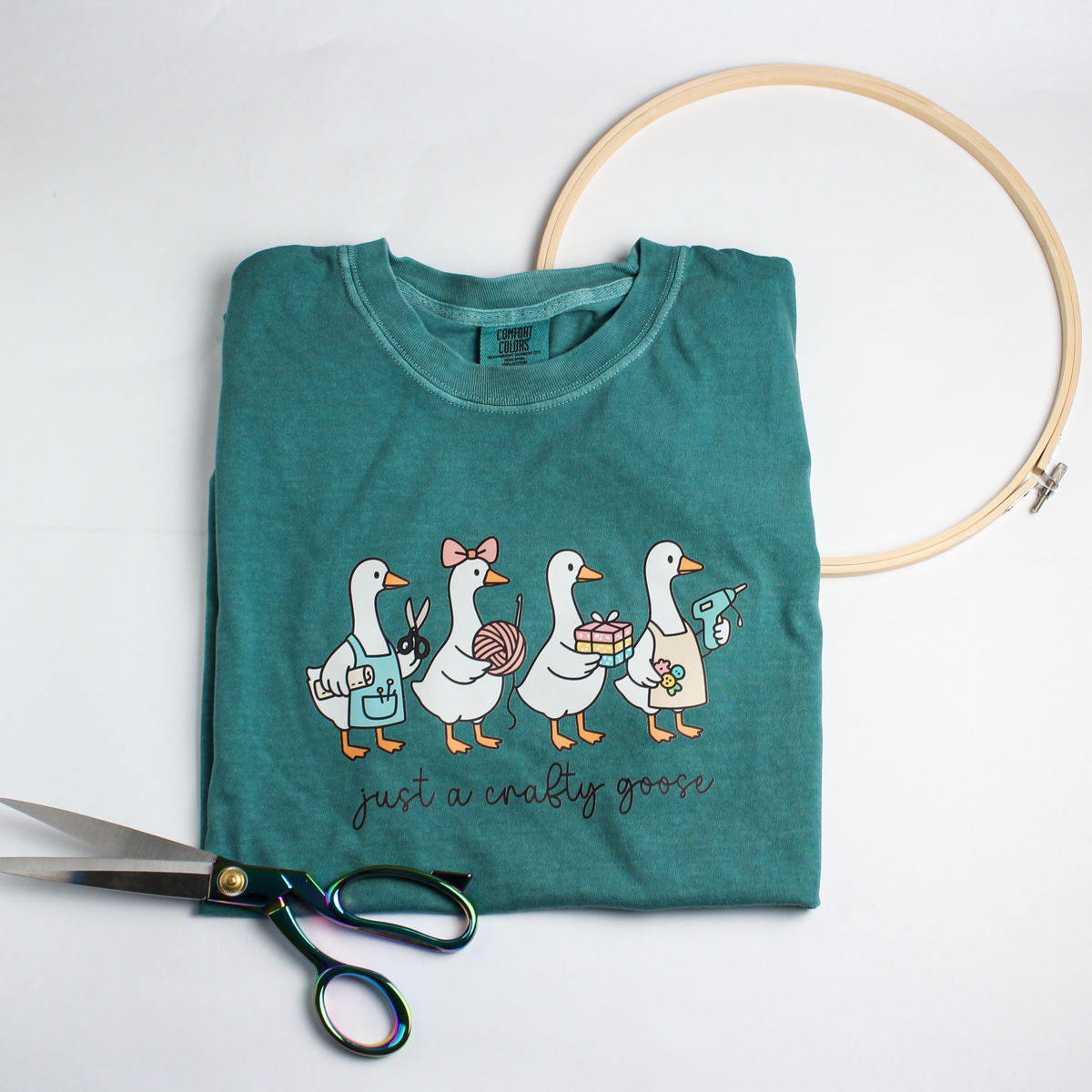 Folded teal Hobby Goose tee with four crafty geese and "just a crafty goose", beside scissors and an embroidery hoop