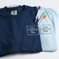 Folded navy blue shirt and Light Blue Shirt with 'Cozy Coloring Club' text, markers, and coloring book on a white background, Front View