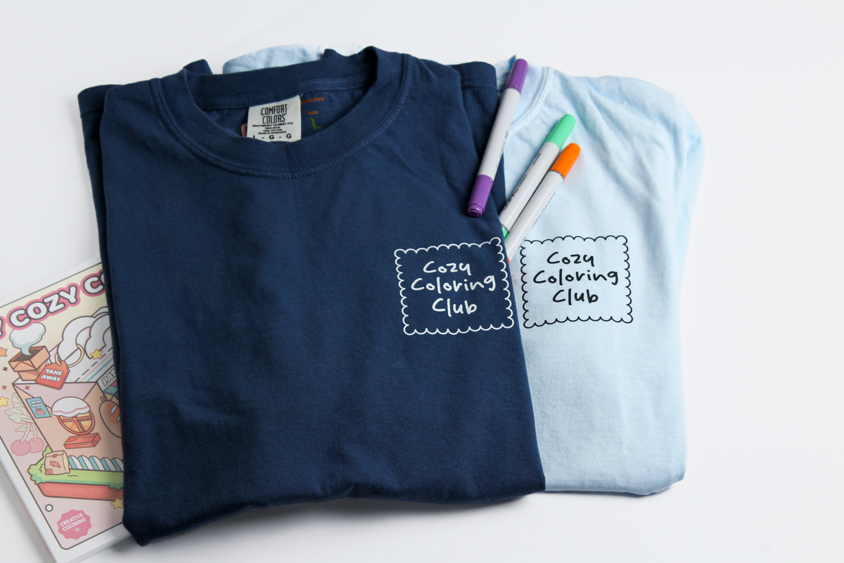 Folded navy blue shirt and Light Blue Shirt with 'Cozy Coloring Club' text, markers, and coloring book on a white background, Front View