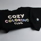 Folded black Cozy Coloring Club crewneck with pastel lettering and a small marker-cup design on the sleeve