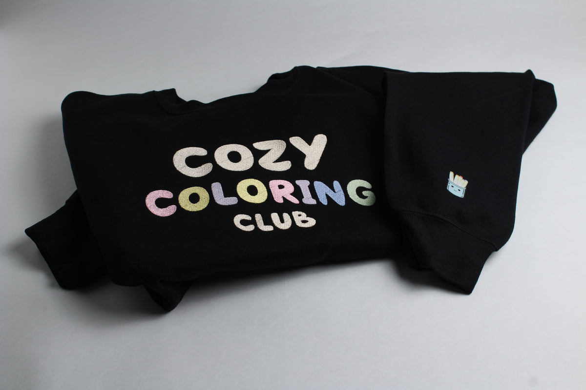 Folded black Cozy Coloring Club crewneck with pastel lettering and a small marker-cup design on the sleeve