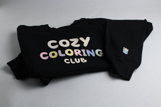 Folded black Cozy Coloring Club crewneck with pastel lettering and a small marker-cup design on the sleeve