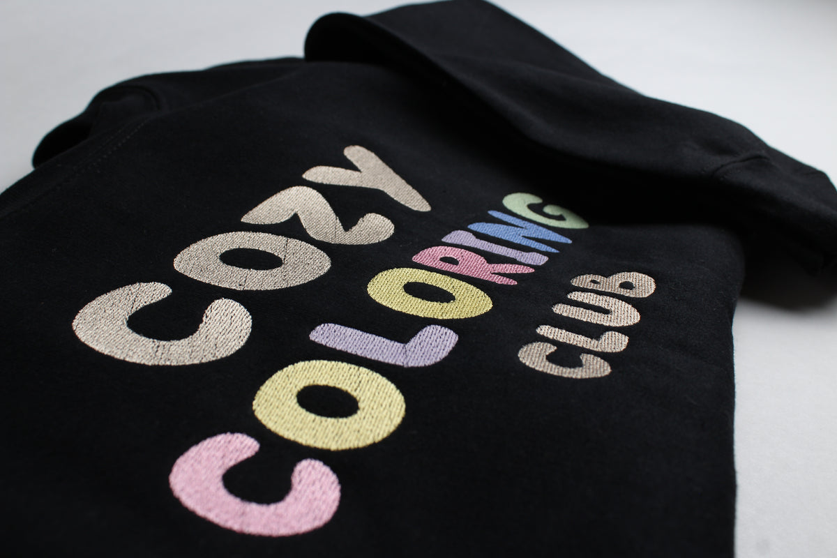 Angled close-up of the pastel Cozy Coloring Club embroidery on a black crewneck
