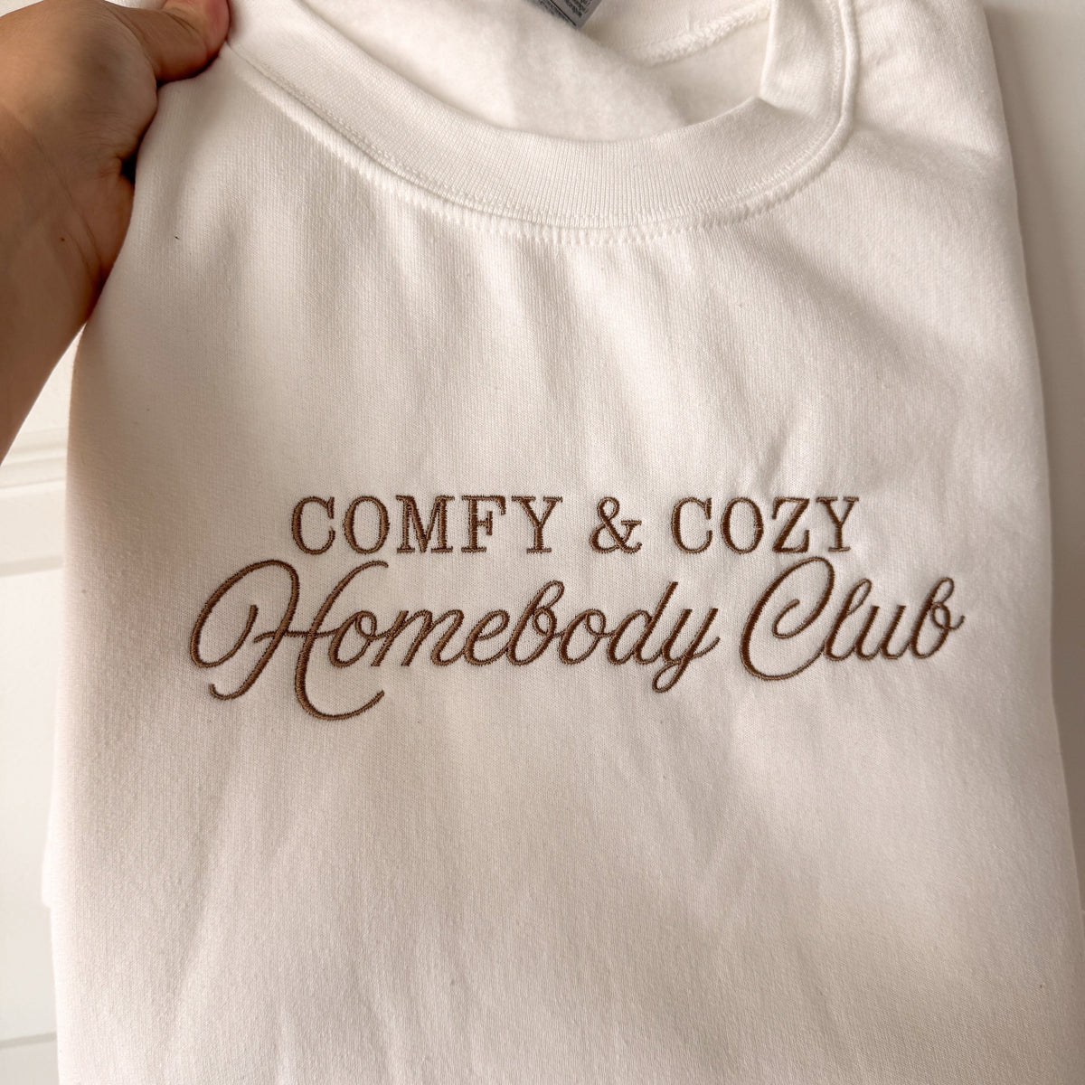White Comfy & Cozy Homebody Club crewneck with brown script embroidery, held up to show the design