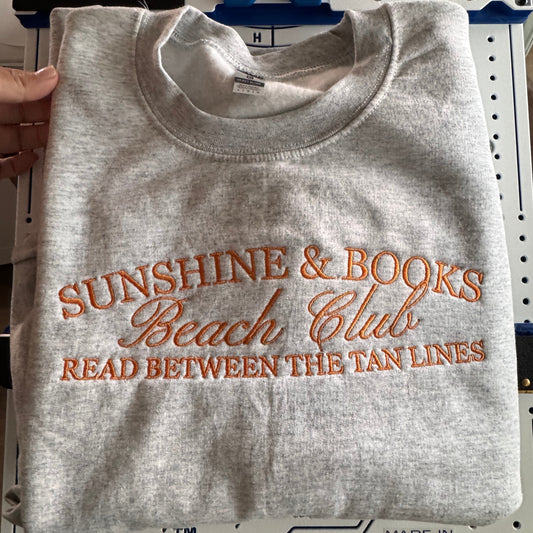 Hand holding a gray crewneck embroidered SUNSHINE & BOOKS Beach Club, Read Between the Tan Lines in orange