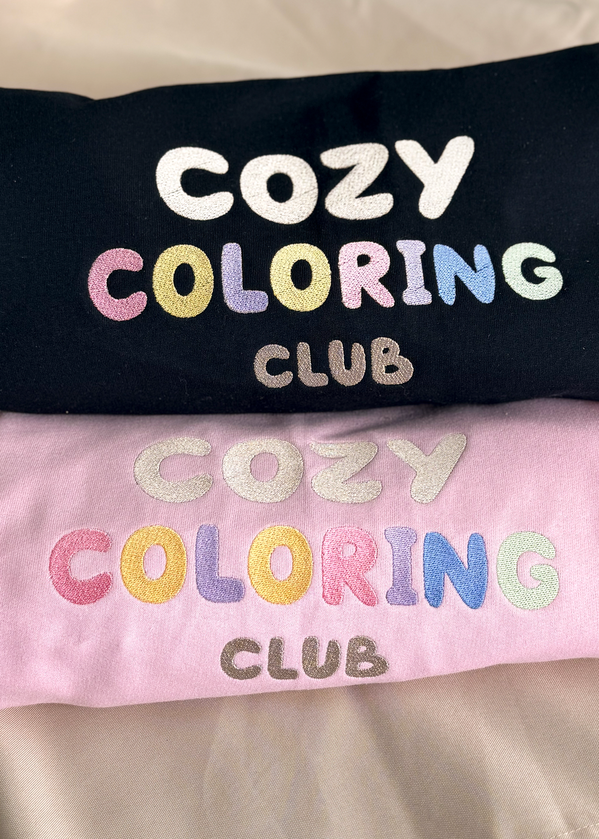 Close-up of the pastel Cozy Coloring Club embroidery on black and pink crewnecks